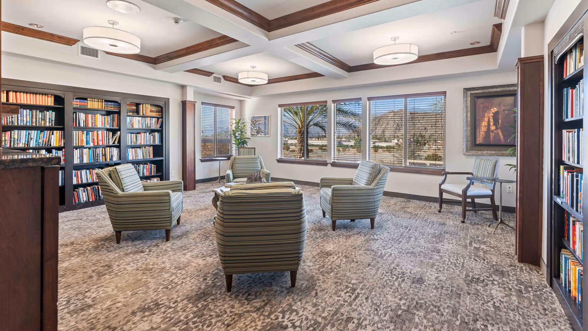 A bright and spacious library or reading room with large windows showing an outdoor view of palm trees and mountains. The room features four striped armchairs arranged around a small wooden table, bookshelves filled with books along the walls, a patterned carpet, and ceiling lights with wooden beams.