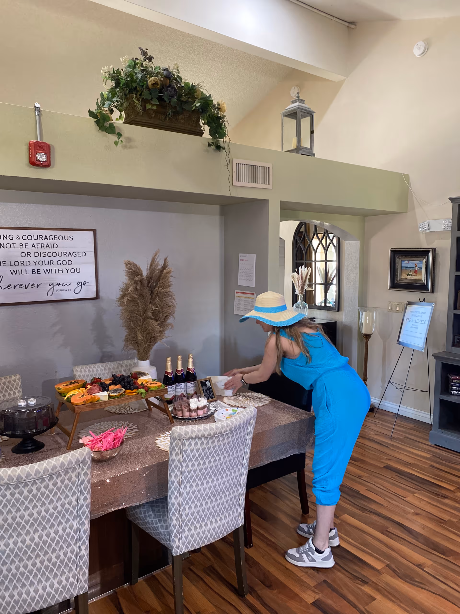 A woman in a blue outfit and wide-brimmed hat is arranging items on a dining table set with a variety of food and drinks in a well-lit room with wooden flooring. The table has a sparkly tablecloth, and there are decorative plants and framed artwork on the walls.