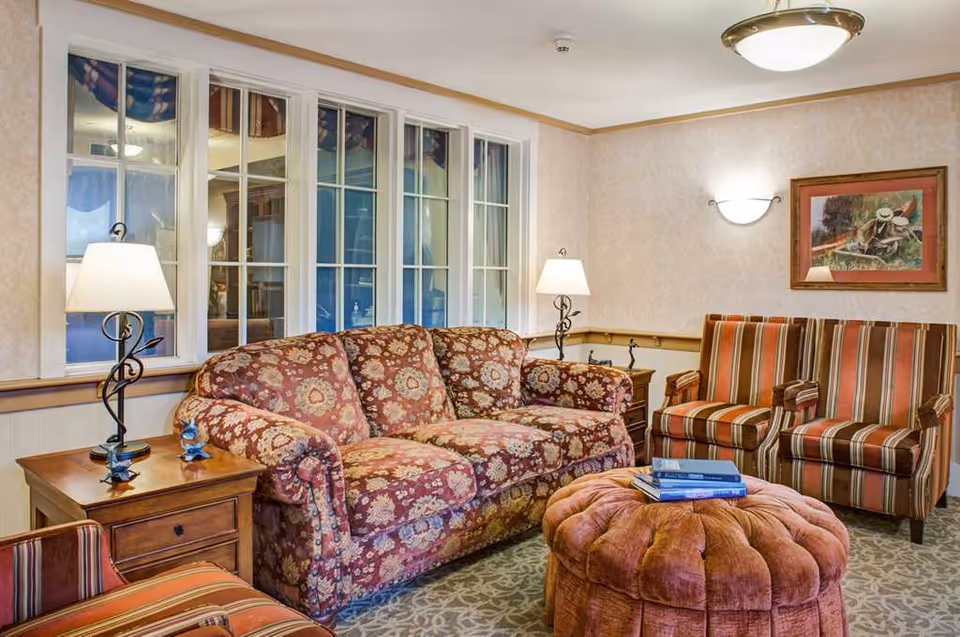 Cozy common living room with a patterned sofa, striped armchairs, a tufted ottoman, side tables and lamps in front of a multi-pane window.