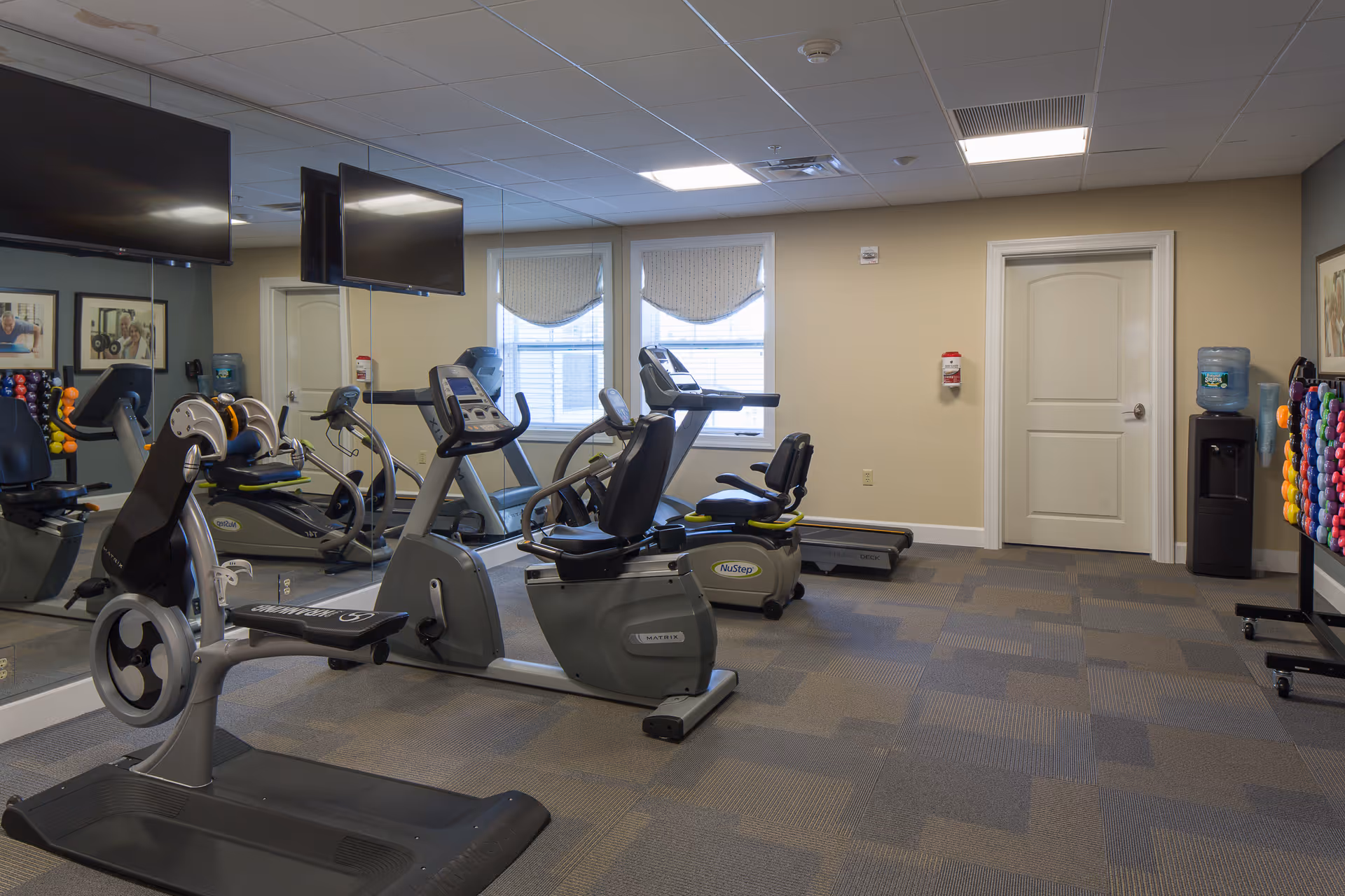 A senior living facility exercise room with various fitness equipment including recumbent stationary bikes, a treadmill, and a weight rack with colorful dumbbells. The room has beige walls, a large mirror on one side, two windows with blinds, a water cooler, and two wall-mounted televisions.