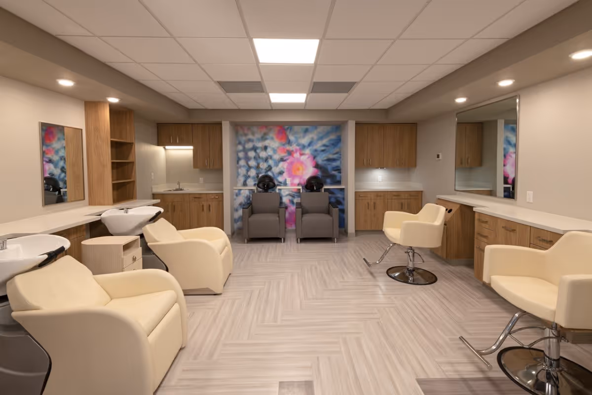 Interior view of a salon area in a senior living facility with cream-colored salon chairs, two hair washing stations, large mirrors on the walls, wooden cabinetry, and a colorful floral mural on the back wall.
