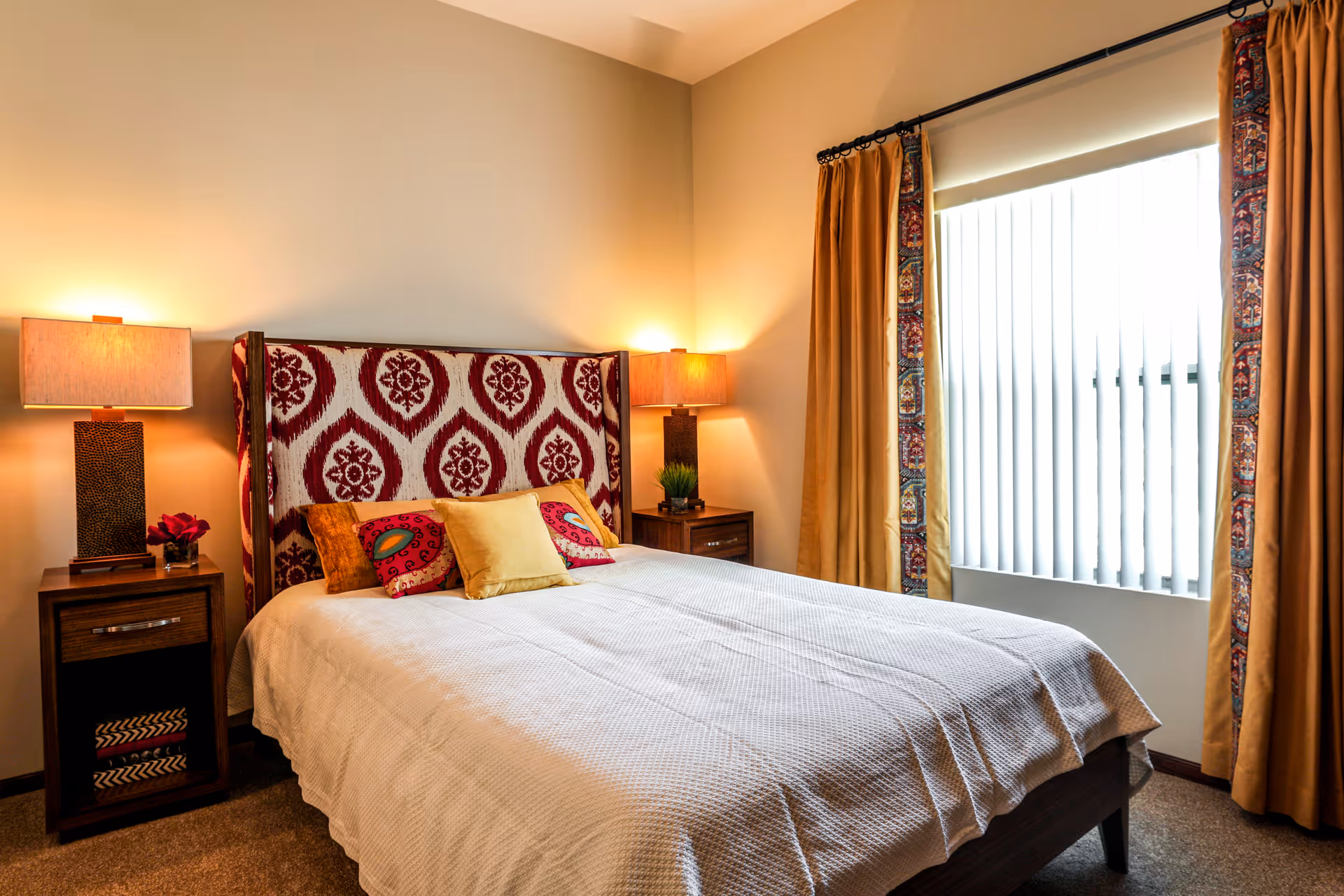 A cozy bedroom with a bed featuring a patterned headboard and several colorful pillows. There are two wooden nightstands on either side of the bed, each with a lamp and decorative items. A window with vertical blinds and mustard-colored curtains is on the right side of the room.