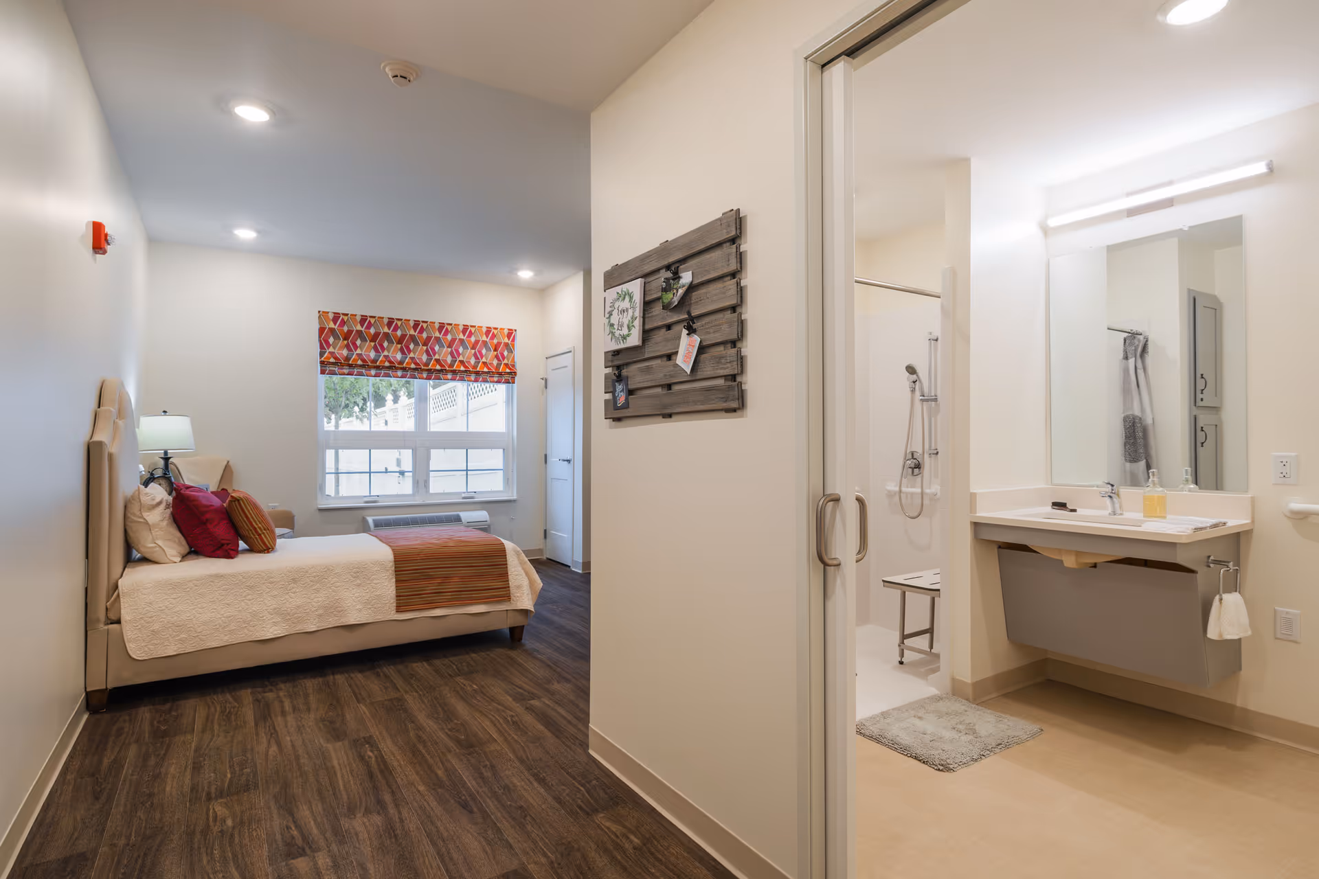 A bright and clean senior living facility room at Heritage Crossing featuring a bed with beige and red pillows and a striped throw blanket. The room has wood flooring, a window with a colorful geometric valance, and a beige armchair next to a lamp. To the right, there is an open doorway leading to a bathroom with a large mirror, sink, and a walk-in shower with a shower chair.