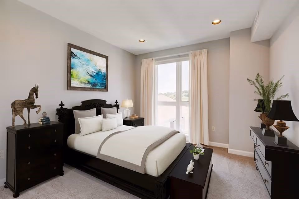 A well-lit bedroom with a large window covered by cream curtains. The room features a dark wooden bed with white and beige bedding, two matching nightstands with lamps, a dresser with a black lamp and a plant, and a chest of drawers with a decorative horse sculpture and books. A colorful abstract painting hangs on the wall above the bed.