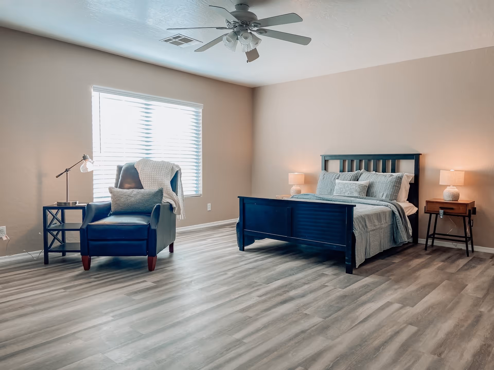 A bedroom with a dark wooden bed frame made with light blue bedding and pillows. There are two wooden nightstands with lamps on each side of the bed. To the left, there is a dark blue armchair with a gray pillow and a white throw blanket, next to a small side table with a lamp. The room has light-colored walls, a large window with blinds, and a ceiling fan with lights. The floor is covered with light gray wood-style flooring.