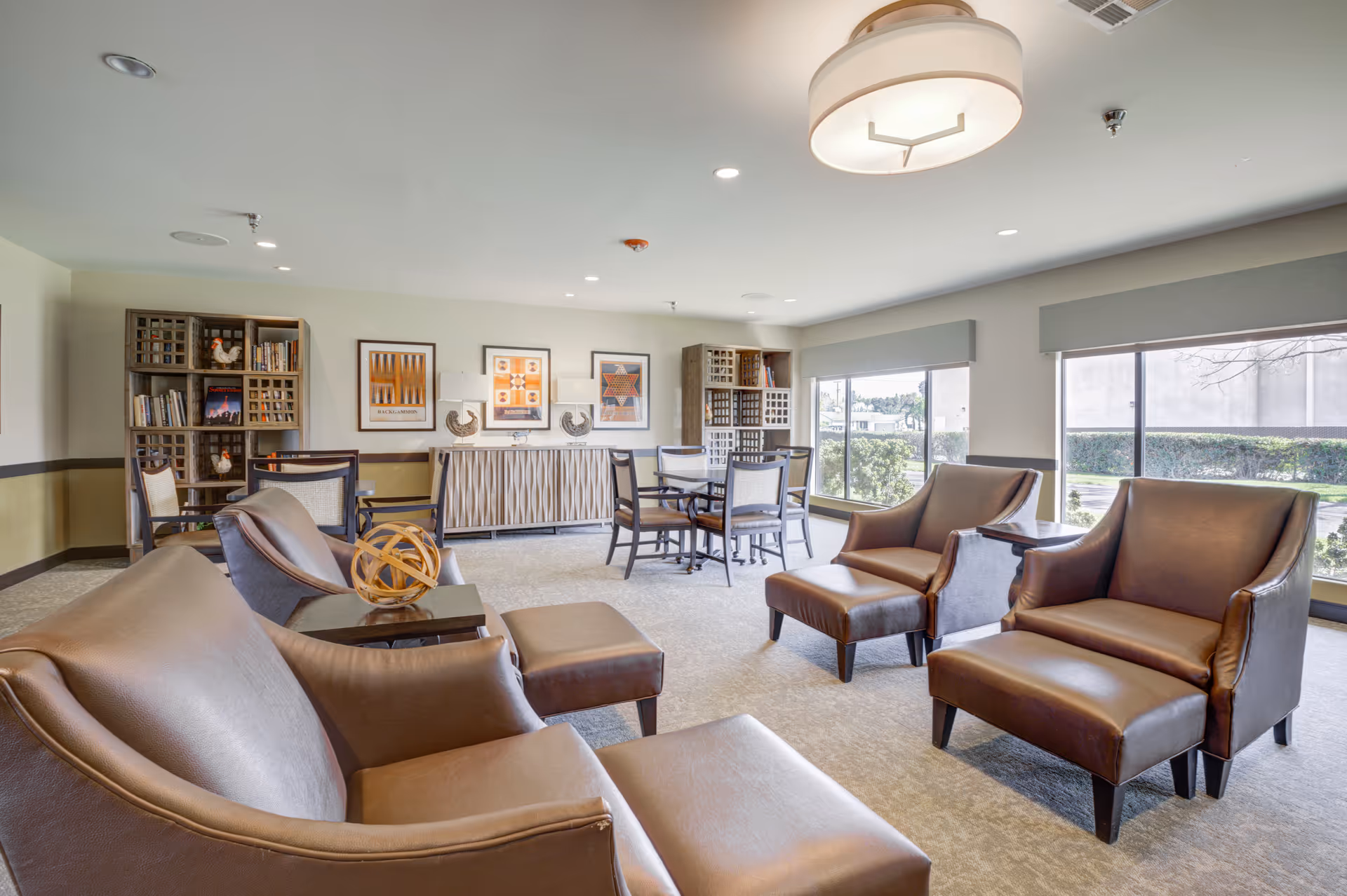 A bright assisted-living common lounge with brown leather armchairs and ottomans, a table, bookshelves, and large windows.