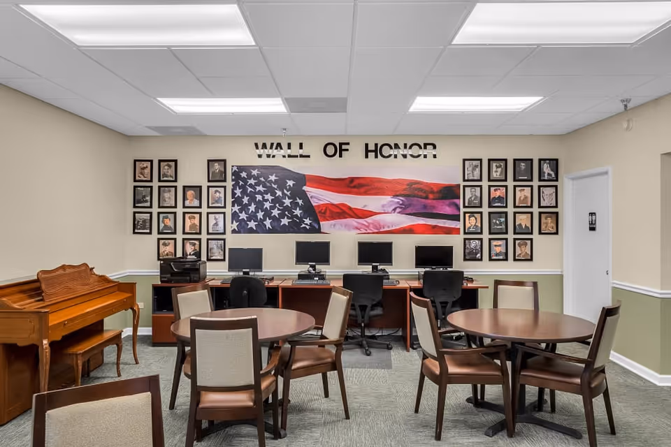 A room with several round tables and chairs, a wooden piano on the left, and a row of computer stations with chairs against the back wall. Above the computers is a large American flag and the words 'WALL OF HONOR' with framed photographs of veterans on either side of the flag.