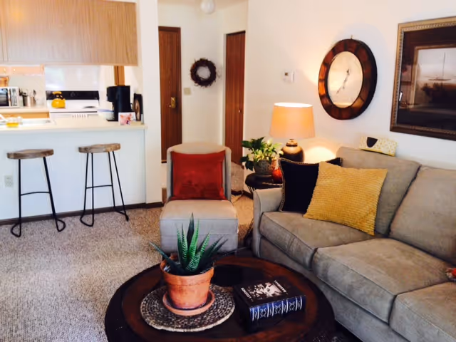Cozy living room with a sofa, armchair, round coffee table with a potted plant, a lamp and wall clock, and a kitchen counter with bar stools in the background.