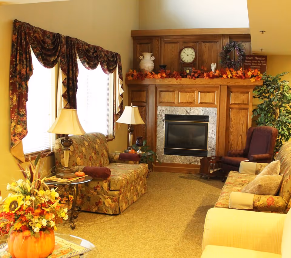 A cozy living room with floral-patterned sofas, a wooden fireplace decorated with autumn leaves, a clock, and vases. There are two table lamps, a large plant, and windows with floral drapes letting in natural light.