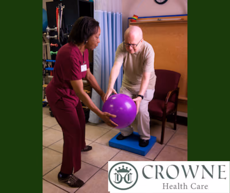 A healthcare worker in burgundy scrubs assists an elderly man who is balancing on a blue foam pad while holding a large purple exercise ball in a therapy room.