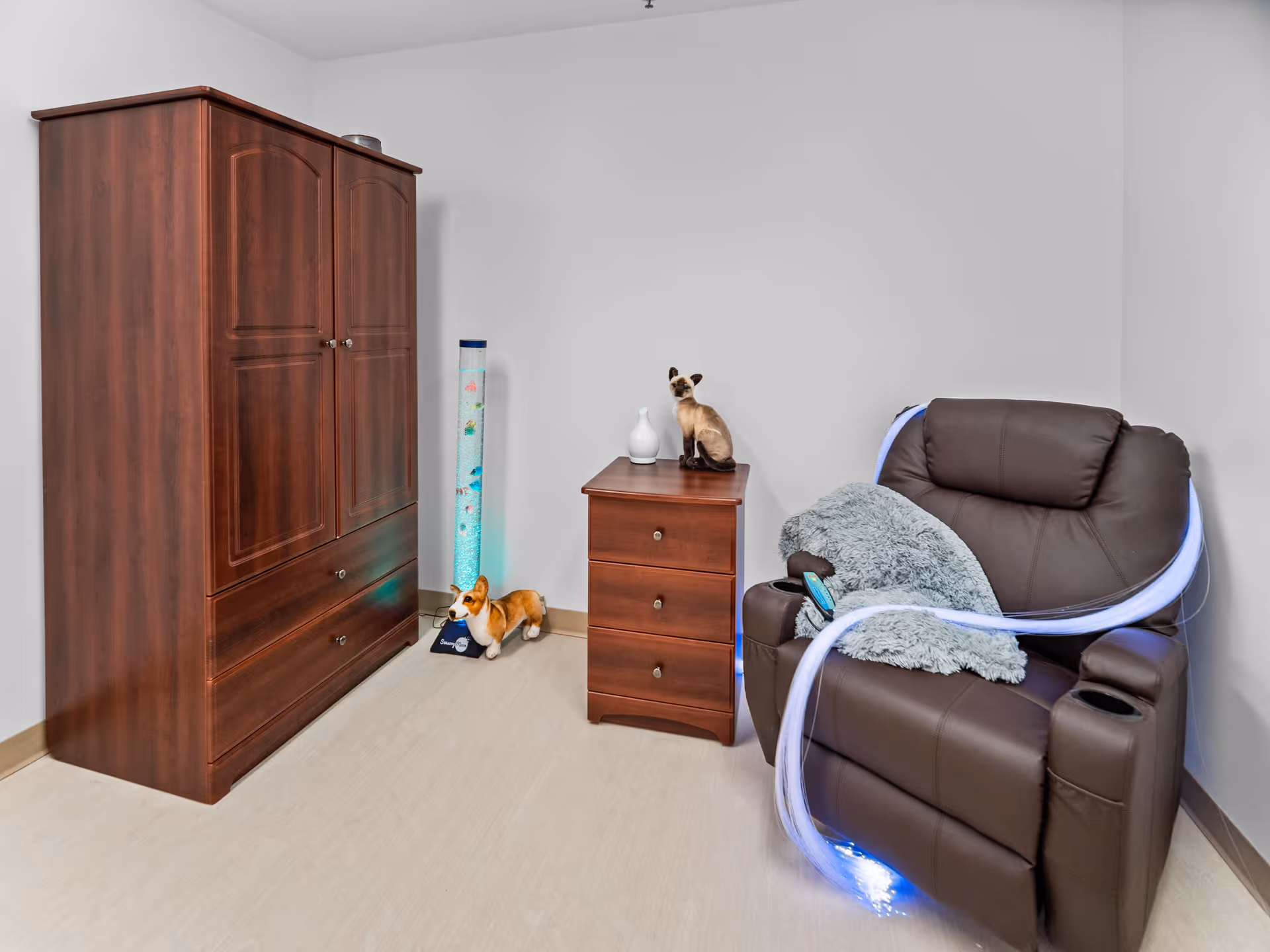 A cozy corner of a room featuring a dark brown recliner chair with a gray fluffy pillow and a glowing light strip draped over it, a small wooden three-drawer nightstand with a decorative cat figurine and a white vase on top, a tall wooden wardrobe, and a small corgi dog figurine on the floor next to a tall cylindrical light with bubbles inside.