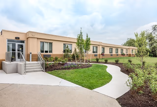 Exterior view of a single-story senior living facility building with beige walls and multiple windows. A concrete pathway curves through a well-maintained lawn and landscaped garden with young trees and shrubs leading to the entrance with stairs and a glass door. The sky is partly cloudy.