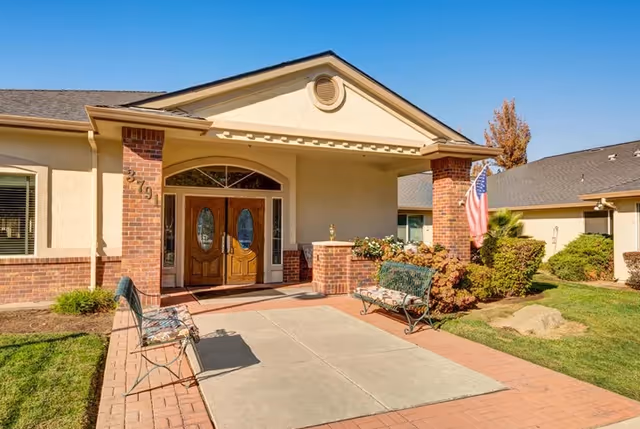 Front entrance of a senior living building with double wooden doors, brick columns, benches, landscaping, and an American flag.