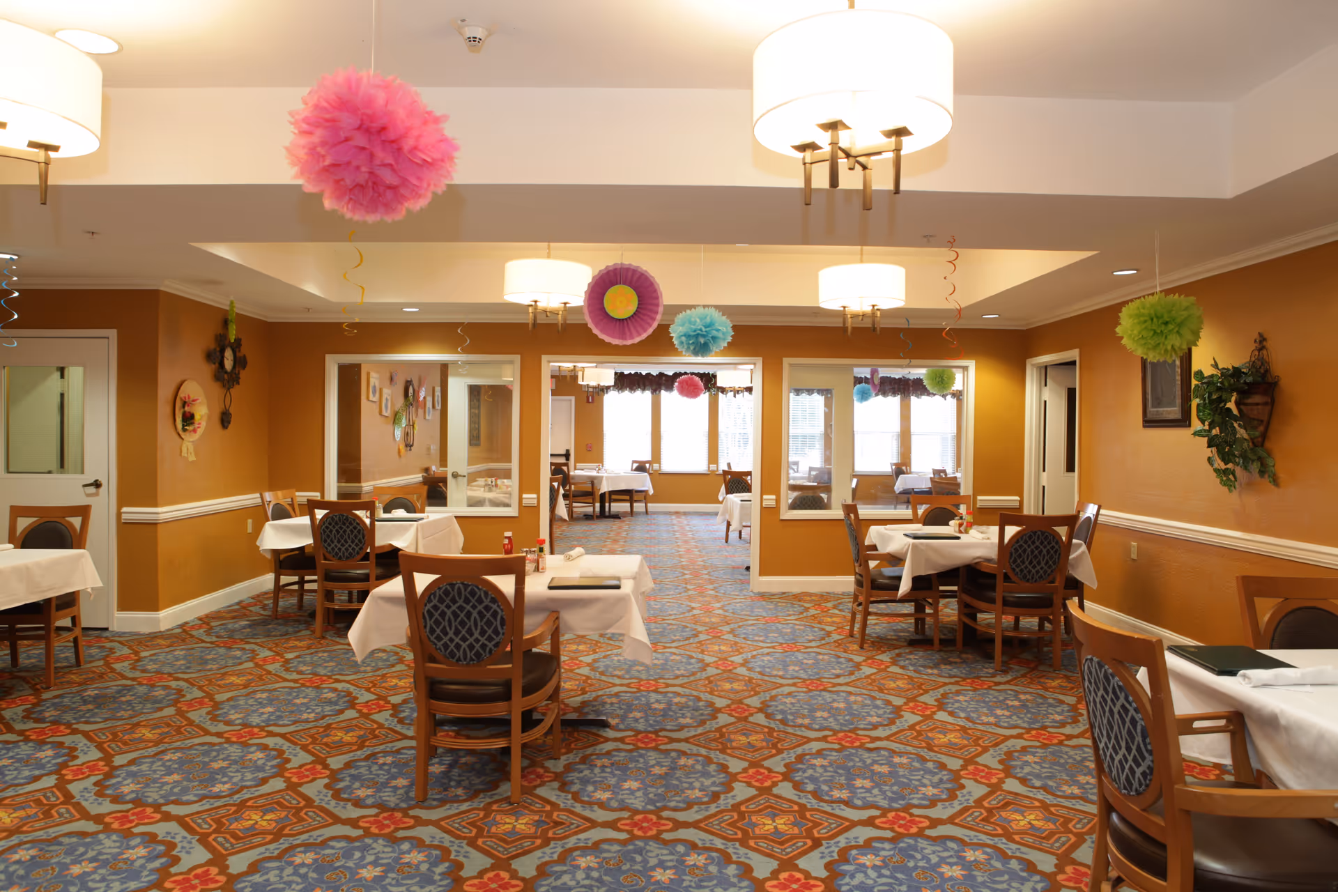 A dining room in a senior living facility with several tables covered with white tablecloths and wooden chairs with patterned cushions. The room has warm orange walls, decorative wall hangings, and colorful hanging paper decorations from the ceiling. Large windows at the far end let in natural light.