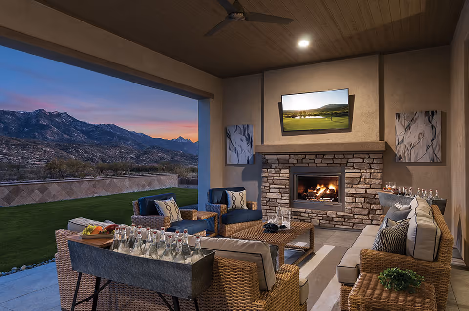 Covered outdoor seating area with wicker furniture including sofas and chairs with cushions, a stone fireplace with a TV mounted above it, and a scenic mountain view at sunset in the background.