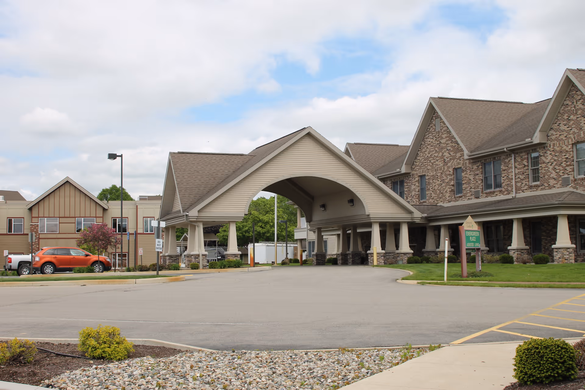 Exterior view of Greencroft Goshen facility showing a large covered entrance with a peaked roof, brick and siding building facade, parked cars, and a sign for Evergreen Place Assisted Living on a grassy area.