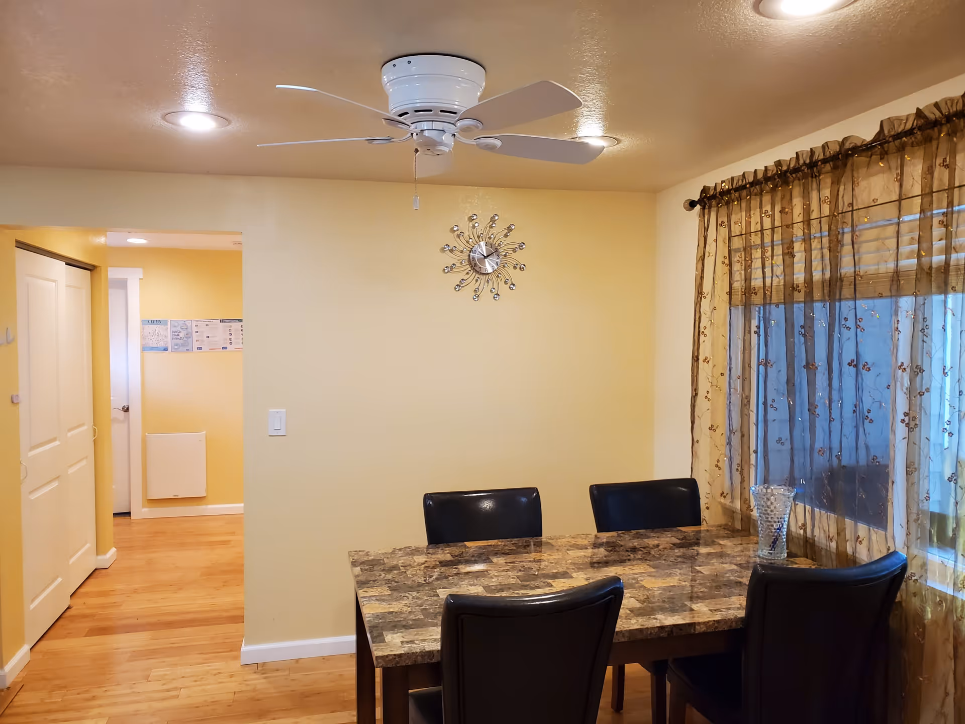 A dining area with a rectangular marble-patterned table surrounded by four black chairs. A decorative clock is mounted on a light yellow wall above the table. A ceiling fan with lights is installed on the ceiling. To the right, a window is covered with sheer brown curtains with a floral pattern. The floor is wooden, and there is an open doorway leading to another room with light yellow walls.