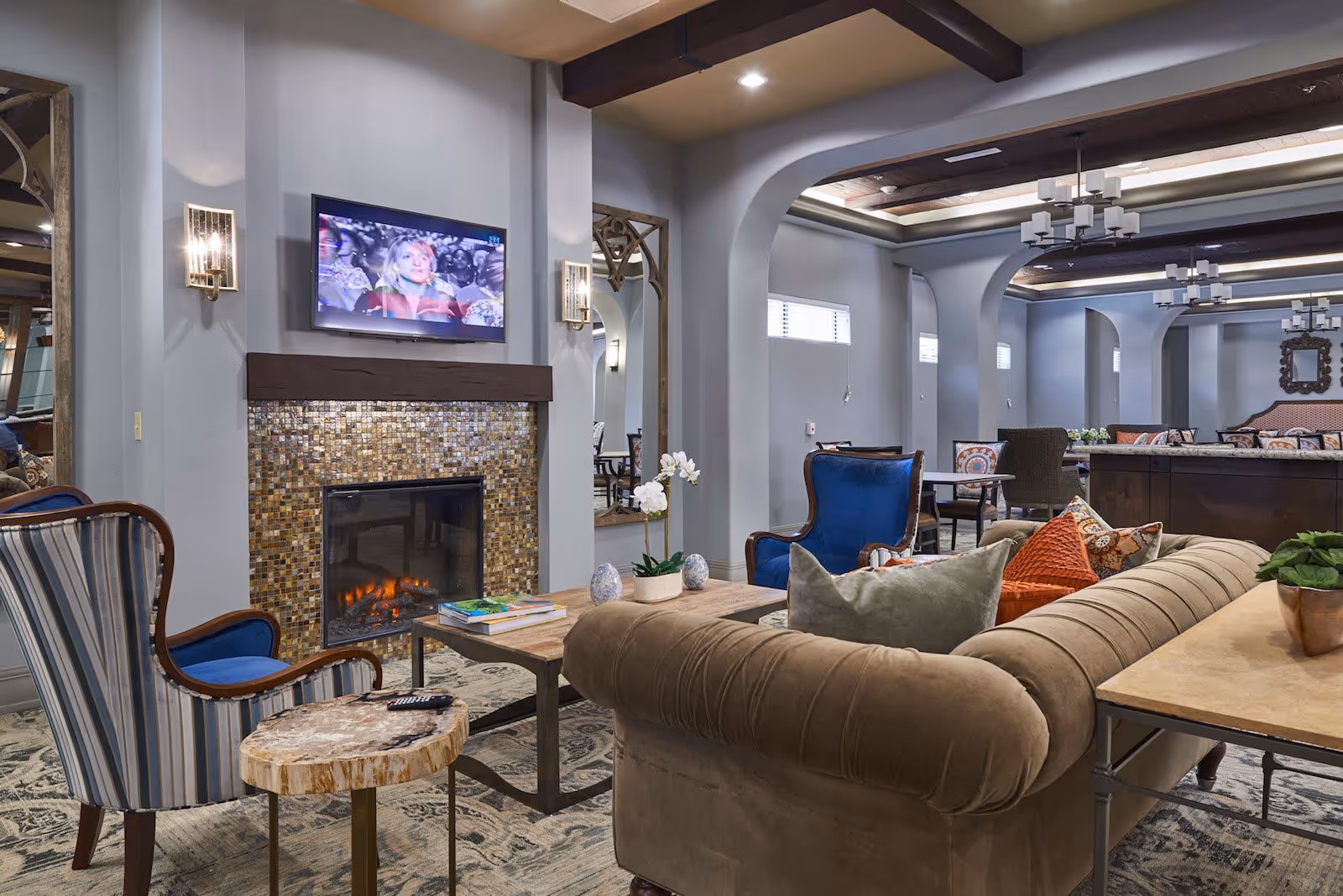 A cozy living room area in an assisted living facility featuring a beige tufted sofa with colorful pillows, a wooden coffee table with books and a small plant, two blue upholstered armchairs, a striped armchair, and a small round wooden side table. The room has a fireplace with a mosaic tile surround and a flat-screen TV mounted above it. The ceiling has exposed wooden beams and modern chandeliers, with large mirrors on the walls and arched doorways leading to other seating areas.