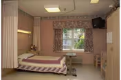A nursing home patient room with a single bed, overbed table, privacy curtain, window with patterned curtains, wall-mounted TV, and wardrobe.