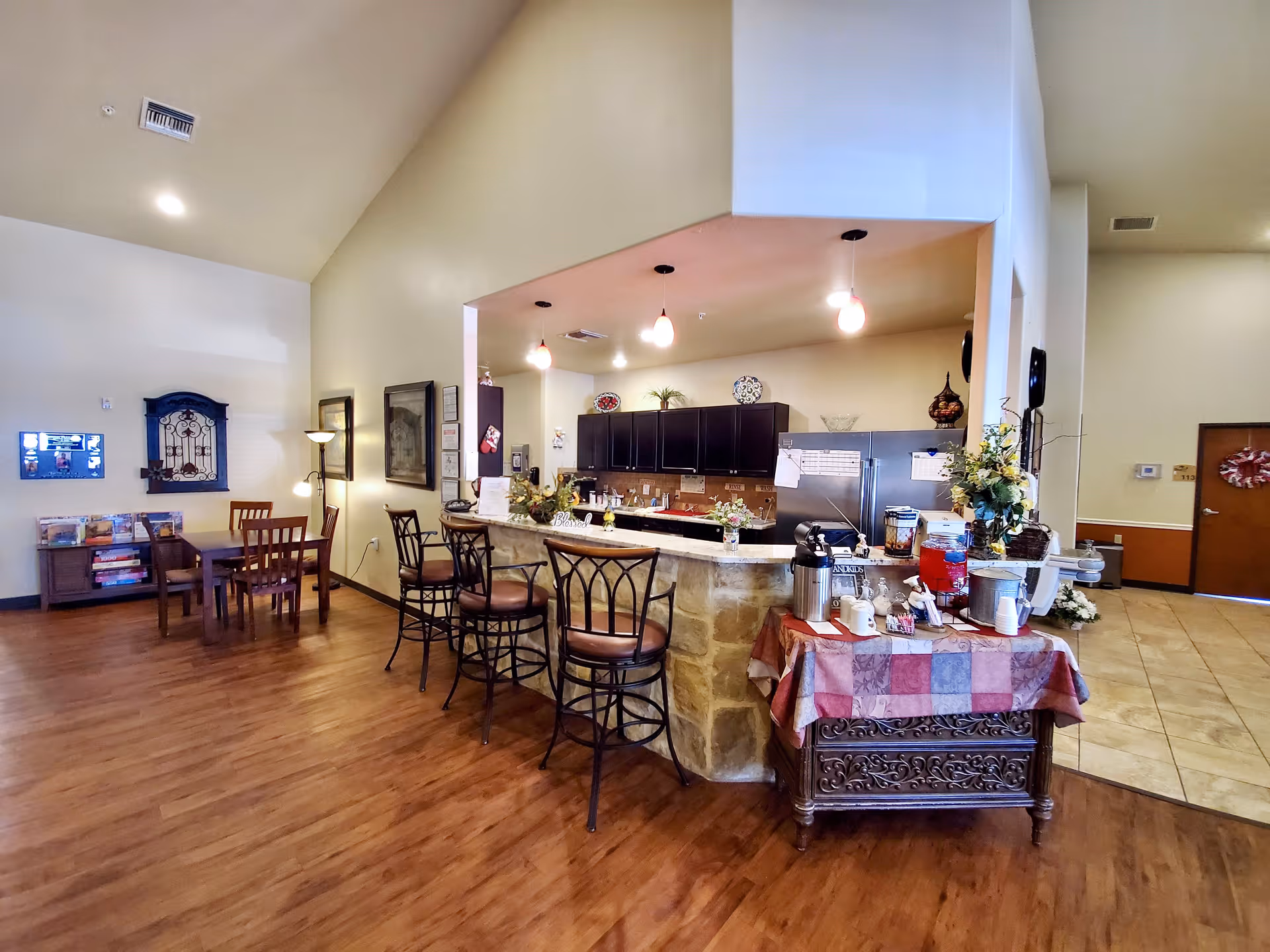 Interior view of a senior living facility showing a kitchen area with a stone and marble counter, three high chairs, and hanging pendant lights. To the right, there is a table covered with a patchwork tablecloth holding coffee and beverage dispensers, cups, and flowers. The floor is wooden, and the walls are painted beige. In the background, there is a small dining area with a table and chairs, a floor lamp, and framed pictures on the wall.