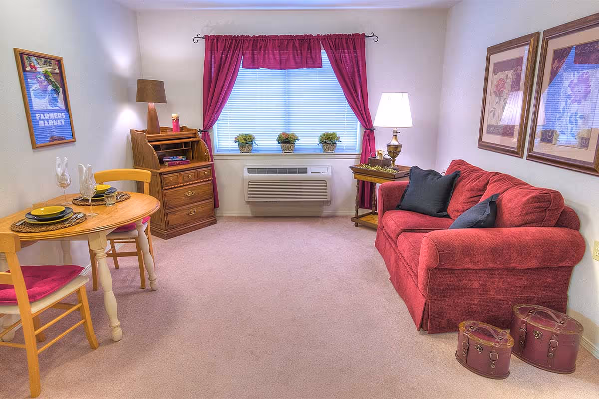 Cozy living area with a red sofa, small round dining table, wooden desk, and a window with burgundy curtains.