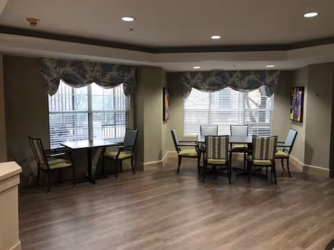 Well-lit communal dining area with round and square tables, chairs, and large windows with patterned valances.