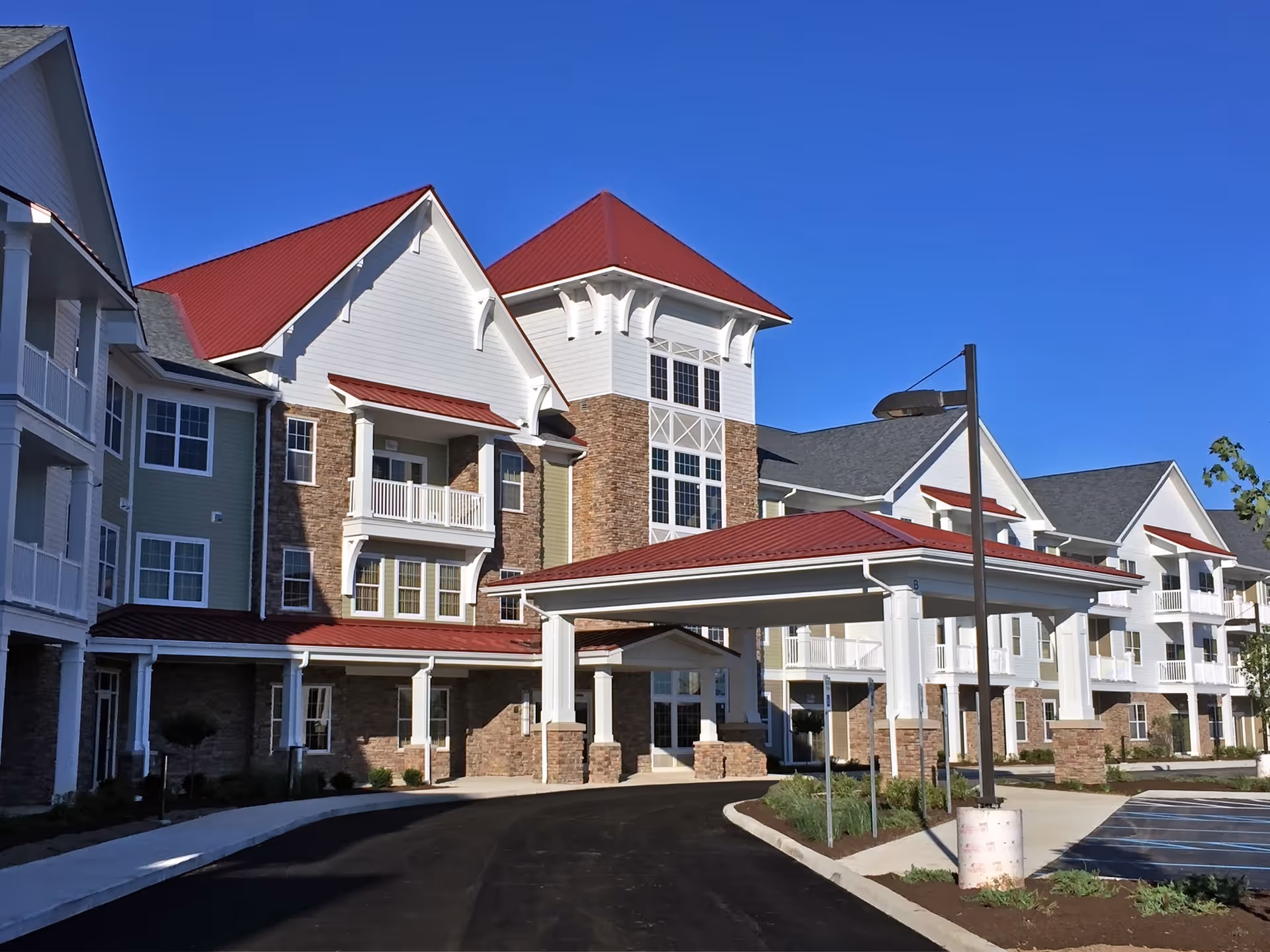 Exterior front entrance of a multi-story senior living building with red roofs, balconies, and a covered porte-cochère under a clear blue sky.
