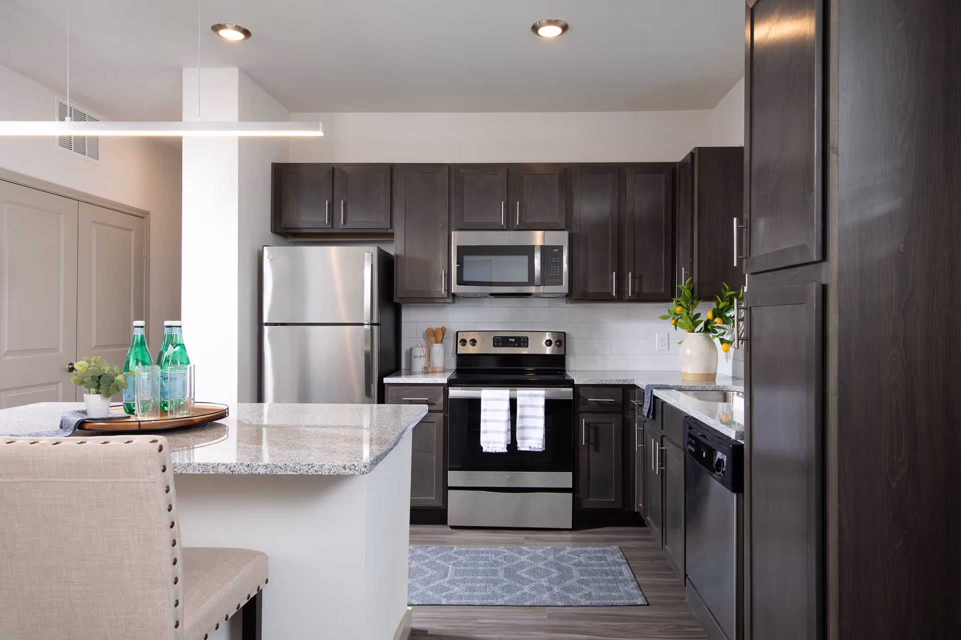 Modern kitchen with dark wood cabinets, stainless steel refrigerator, oven, microwave, and dishwasher. Granite countertops with a small island featuring a tray with green glass bottles and glasses. A beige upholstered chair is partially visible in the foreground.