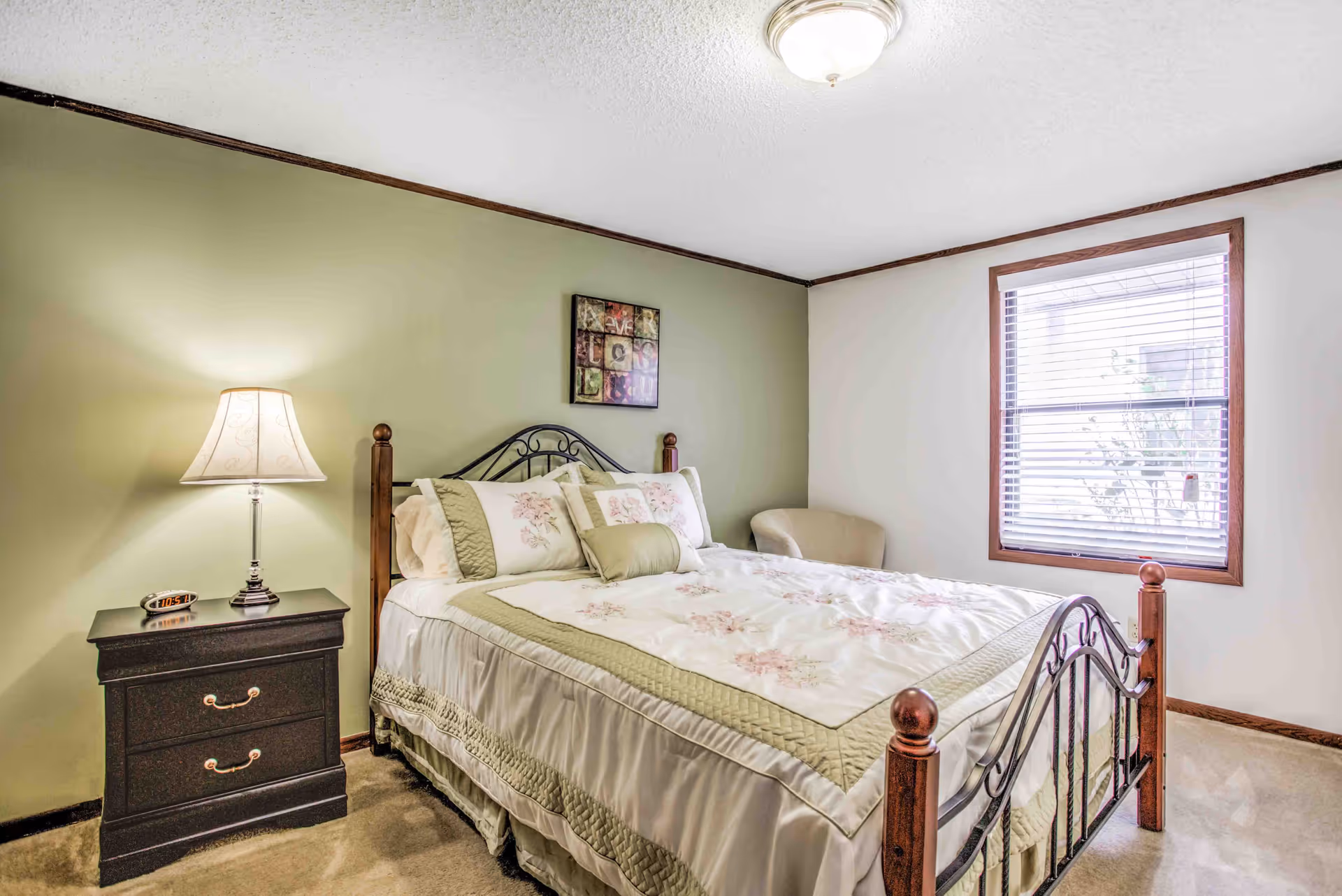 A bedroom with a bed featuring a metal and wood frame, floral bedding, and multiple pillows. There is a nightstand with a lamp and a digital clock on it to the left of the bed. A beige armchair is placed near a window with blinds. The walls are painted light green and white, and there is a framed artwork hanging above the bed.