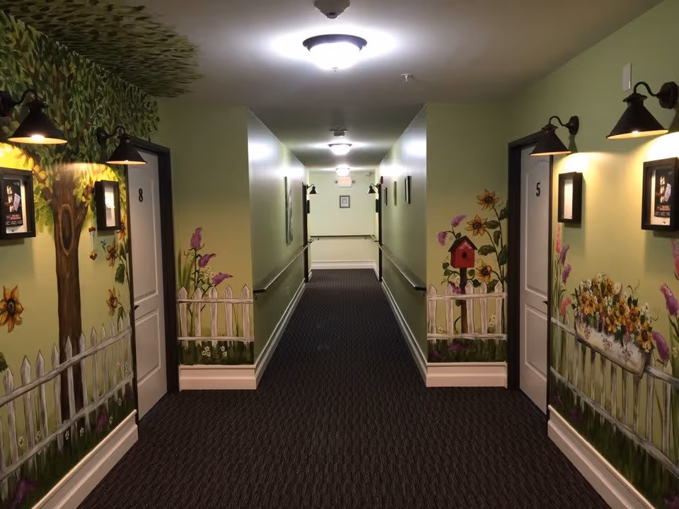 A well-lit hallway in a memory care facility with doors numbered 5 and 8 on either side. The walls are painted light green and decorated with colorful murals of trees, flowers, a white picket fence, and a birdhouse. Wall-mounted lights illuminate framed pictures next to each door. The floor is carpeted in a dark pattern, and handrails run along both sides of the hallway.