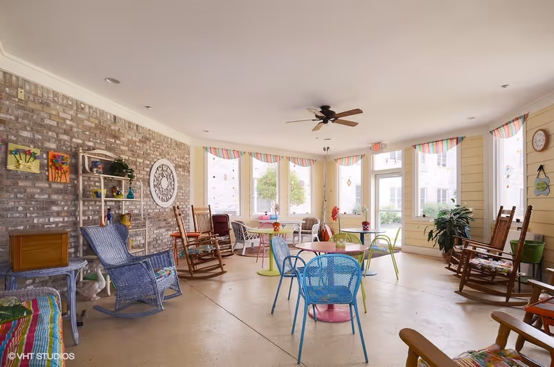 A bright and airy common area with multiple colorful chairs and tables, rocking chairs, large windows with striped valances, a ceiling fan, and decorative wall art. The room has a brick accent wall and light yellow walls, creating a welcoming and cheerful atmosphere.