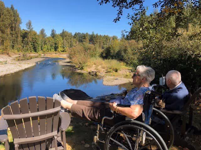 Two elderly individuals in wheelchairs sitting outdoors near a calm river surrounded by trees and greenery under a clear blue sky.