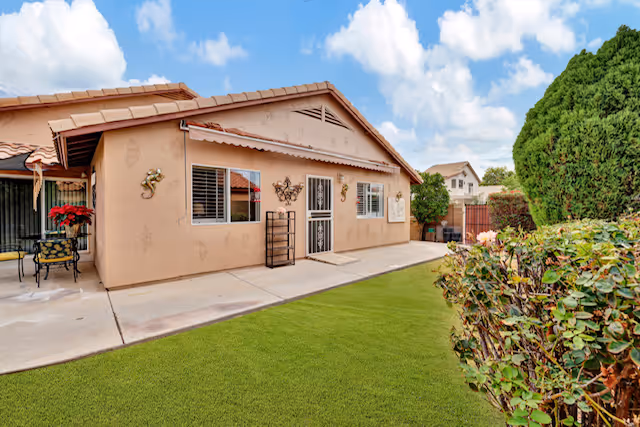 Exterior view of a single-story building with a beige stucco finish and a tiled roof, featuring a patio area with chairs and a table, decorative wall hangings, and a well-maintained green lawn bordered by bushes under a partly cloudy blue sky.