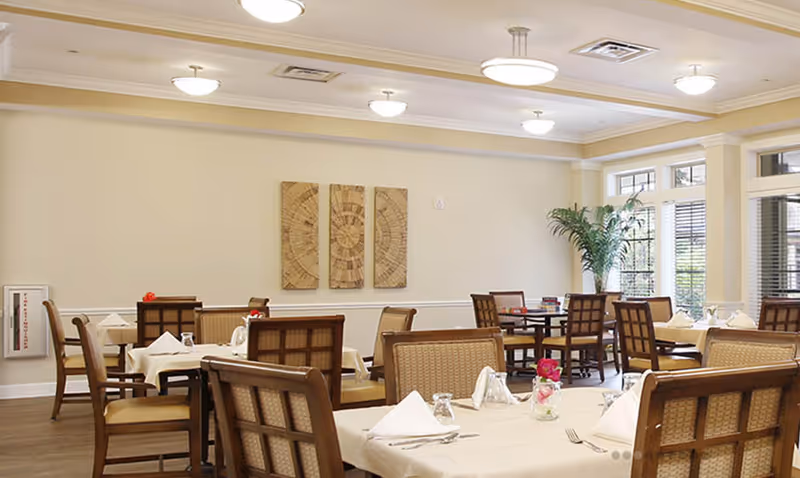 A bright dining room with multiple tables covered in white tablecloths, each set with napkins, glasses, and silverware. The room has wooden chairs with woven backs, beige walls, ceiling lights, and large windows letting in natural light. There is a green potted plant in the corner and three wooden wall art pieces on the wall.