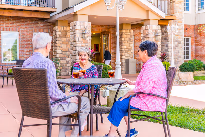 Three older adults sitting at a patio table outside a senior living building, smiling and enjoying drinks.