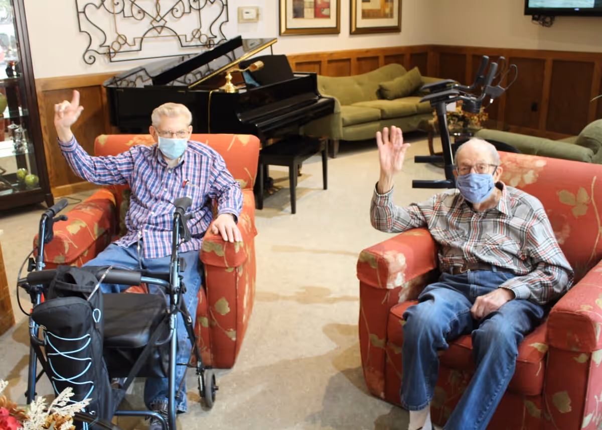 Two elderly men wearing face masks sit in red armchairs in a communal living room, waving with a piano and exercise equipment behind them.