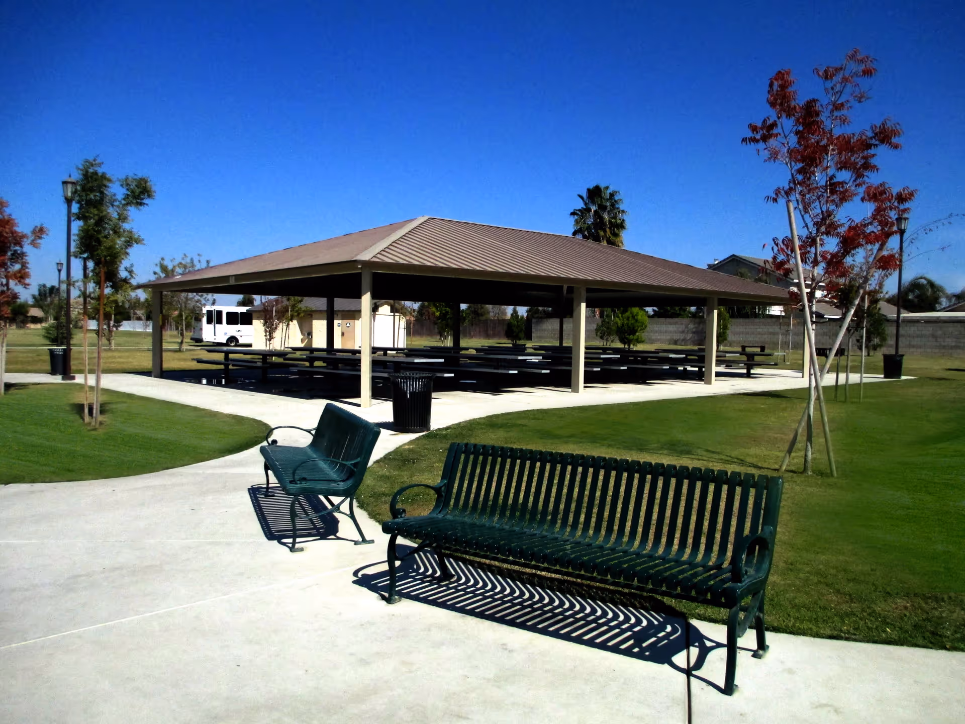 Outdoor park area with green metal benches on a concrete walkway, a covered pavilion with picnic tables, green grass, young trees, and a clear blue sky.