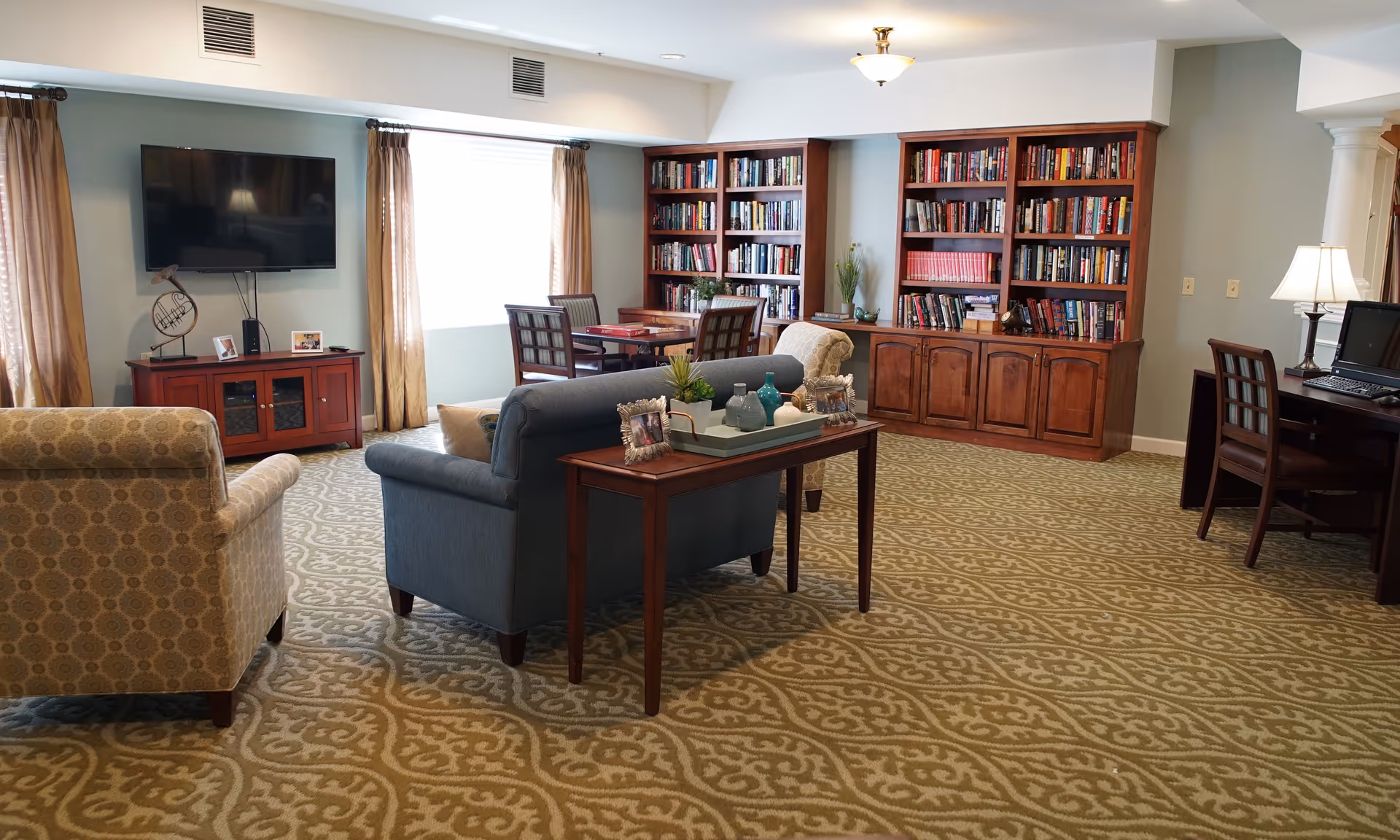 A cozy living room area in a senior living facility with a patterned carpet, a blue sofa, two beige armchairs, a wooden console table with decorative items and photo frames, a large flat-screen TV mounted on the wall above a wooden cabinet, a window with curtains, a wooden bookshelf filled with books, a small table with four chairs, and a desk with a computer and lamp.