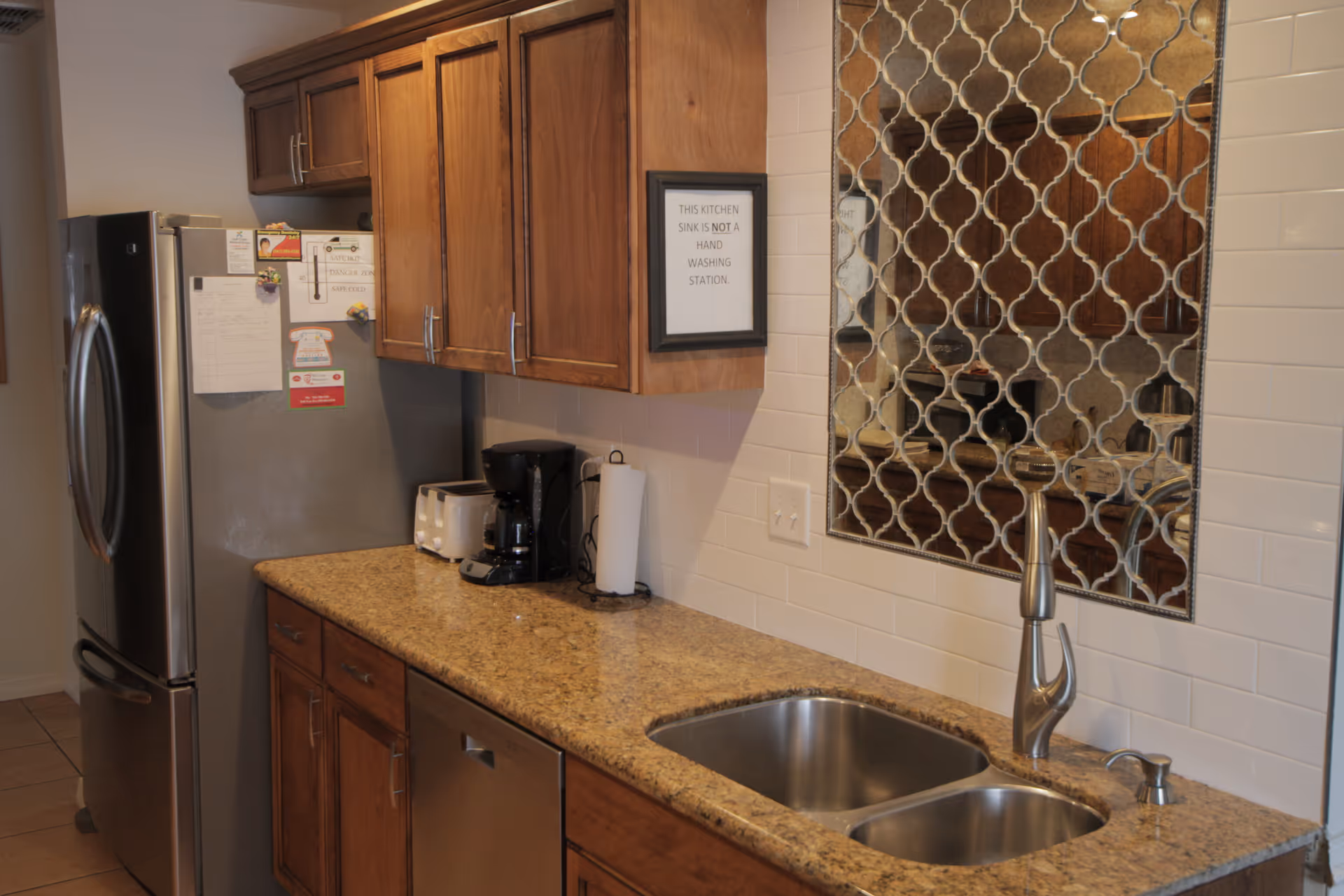A kitchen area with wooden cabinets, a granite countertop, a double stainless steel sink with a modern faucet, a coffee maker, a toaster, a paper towel holder, and a stainless steel refrigerator. There is a decorative mirror with a patterned frame on the wall above the sink and a sign that reads 'THIS KITCHEN SINK IS NOT A HAND WASHING STATION.'