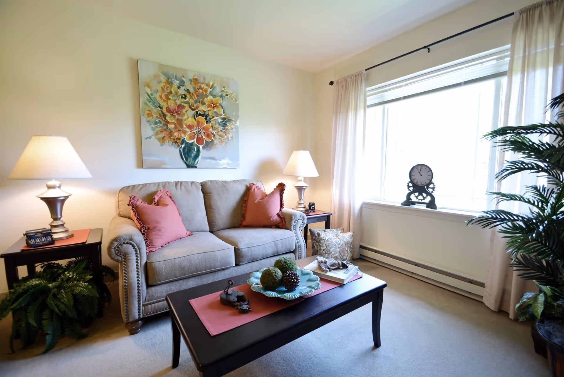 Comfortable living room with a beige upholstered sofa flanked by lamps, a coffee table with decorative items, wall art, plants and a large window.