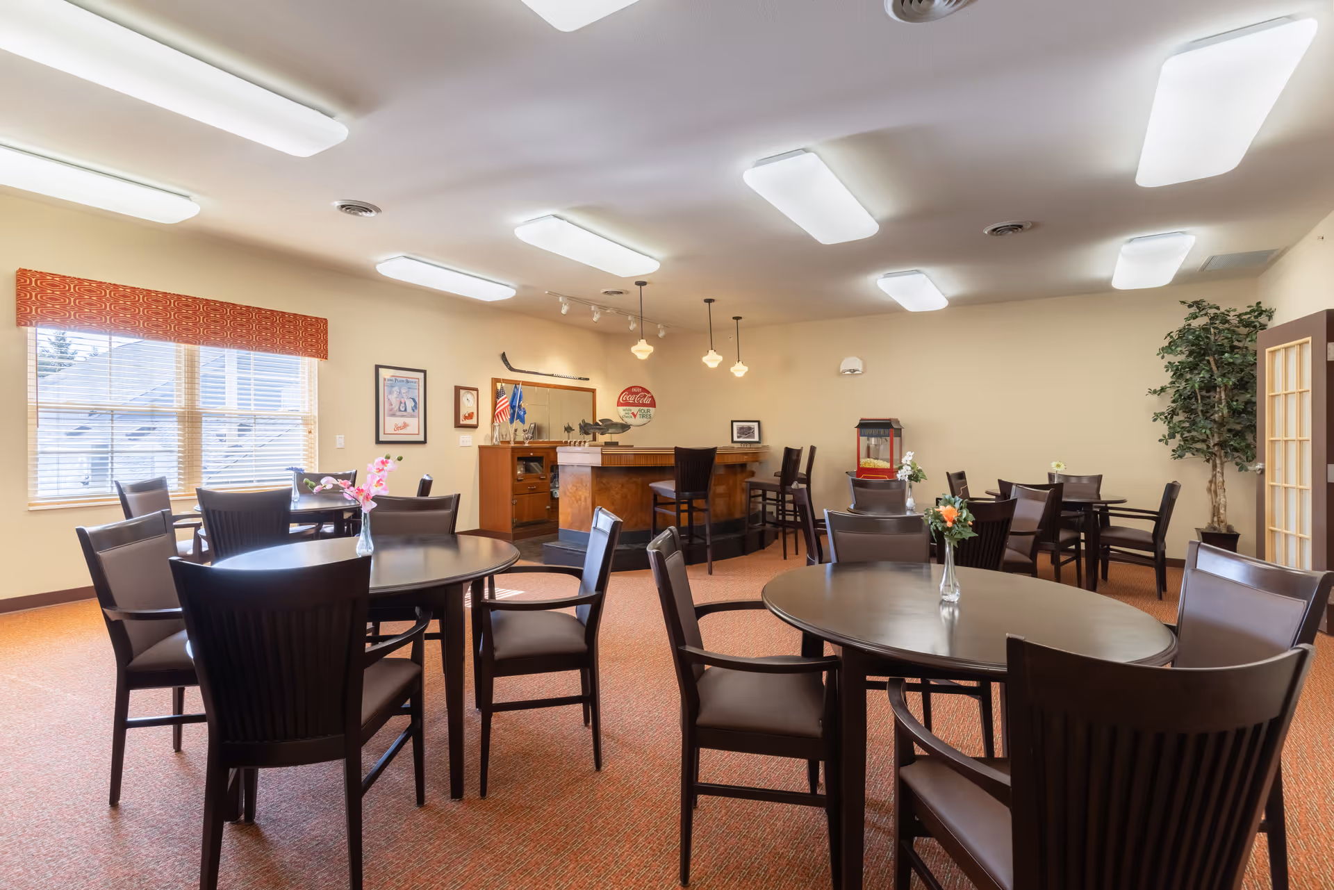 A bright dining room with several round and square tables surrounded by dark wooden chairs. Each table has a small vase with flowers. There is a window with blinds and a red valance on the left wall. The back wall features a small bar area with stools, vintage Coca-Cola signage, and framed pictures. A popcorn machine and a tall potted plant are also visible in the room.