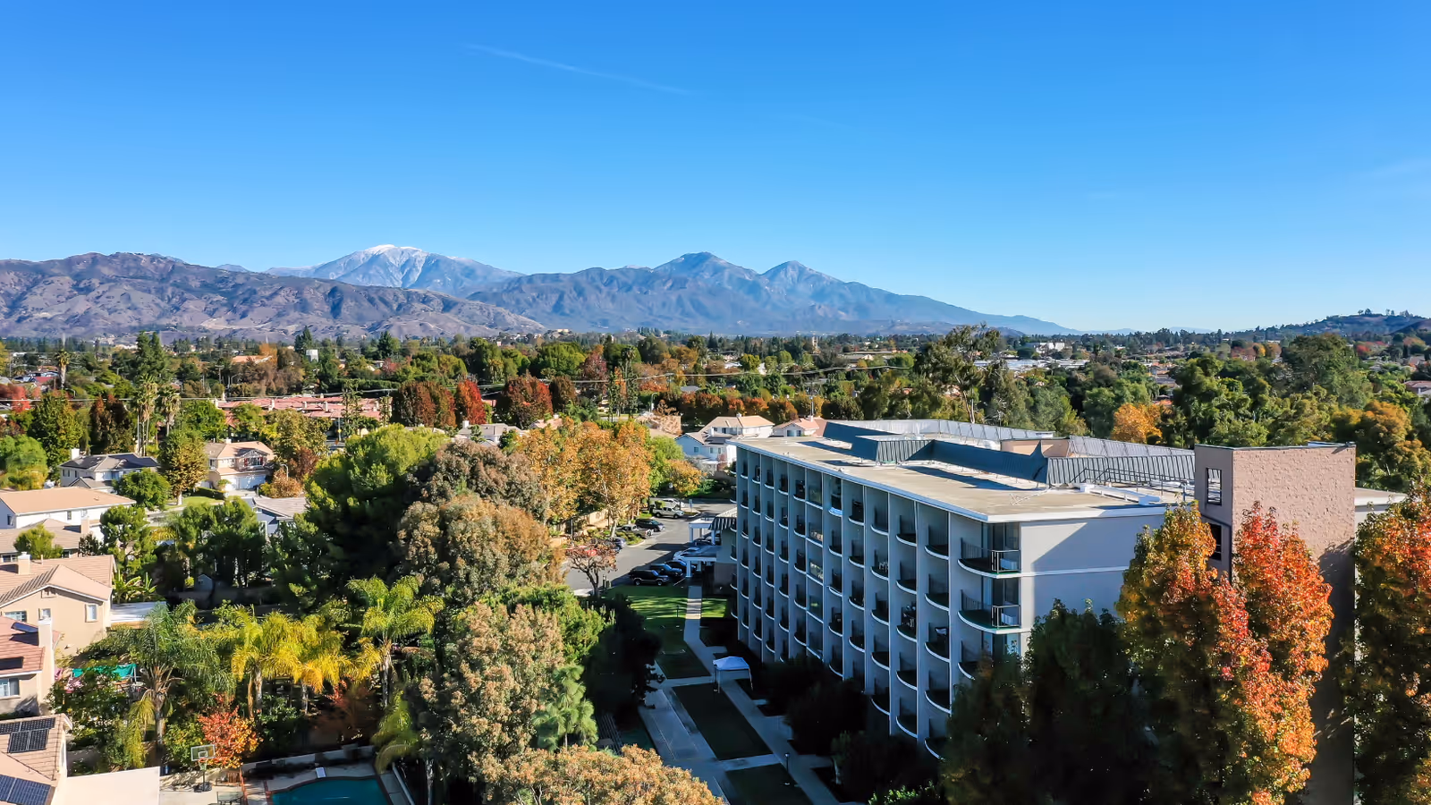 Aerial view of a multi-story senior living building with balconies surrounded by trees and neighborhood homes, with mountains in the distance.