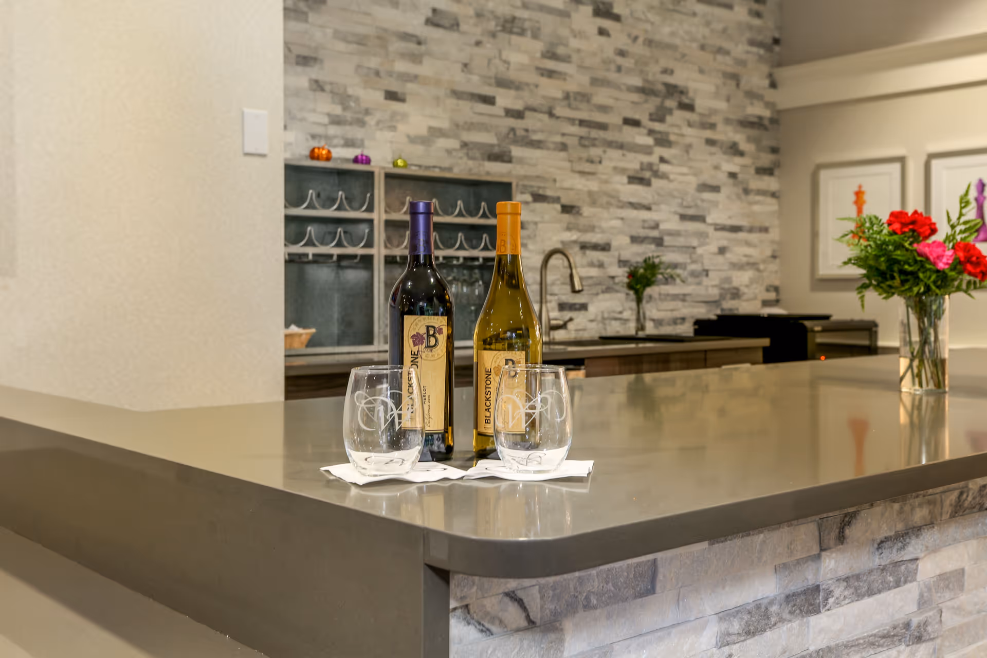 A modern kitchen countertop with two bottles of wine and two empty wine glasses on white napkins. The background features a stone tile backsplash, a sink with a faucet, a wine rack, and a vase with red and pink flowers.