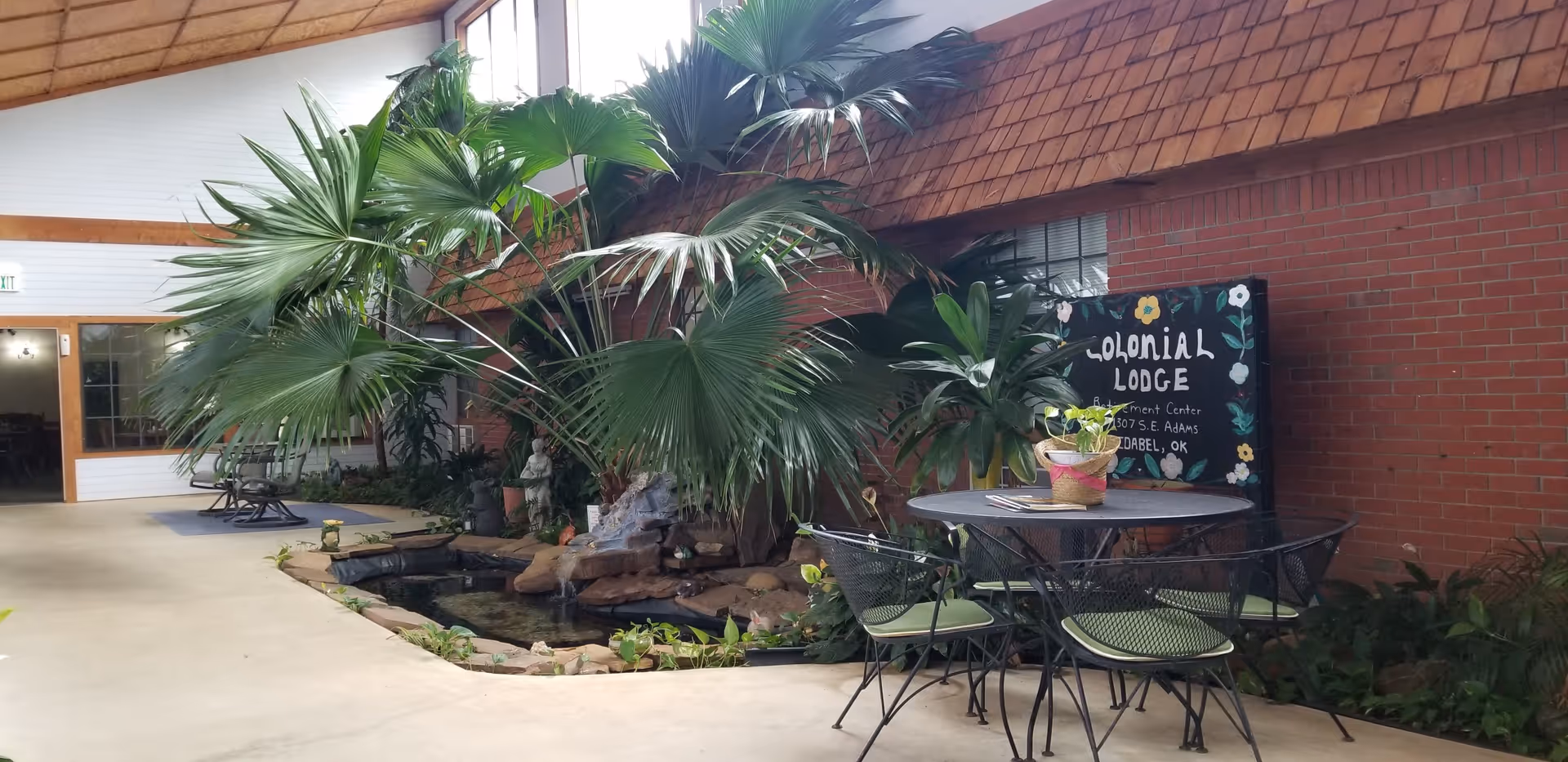 Indoor seating area with a round metal table and four chairs with green cushions next to a small indoor pond with rocks and a water feature. Large green tropical plants surround the pond. A brick wall with a sign reading 'Colonial Lodge Retirement Center 1307 S.E. Adams Idabel, OK' is visible behind the plants. The ceiling is wooden with skylights allowing natural light.
