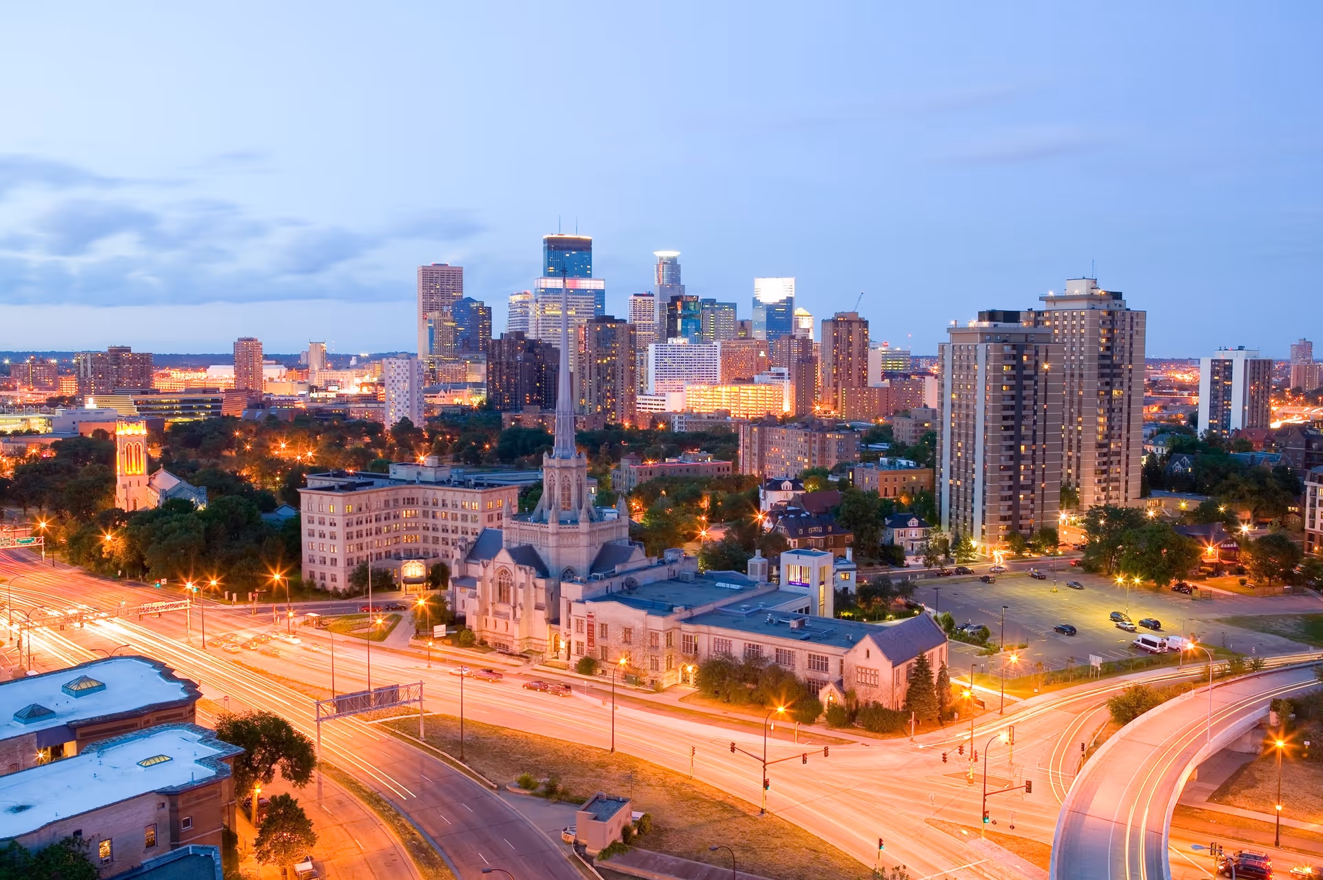 A cityscape at dusk showing a busy highway with light trails from vehicles, a prominent church with a tall steeple in the foreground, and a skyline of tall buildings and skyscrapers in the background under a clear sky.