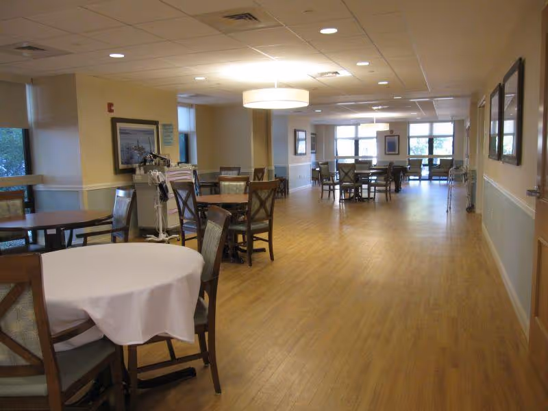 Interior view of a spacious dining area with multiple wooden tables and chairs arranged neatly. The floor is wooden, and the ceiling has recessed lighting and a large circular light fixture. Windows along the left and far end walls allow natural light to enter. The walls are decorated with framed pictures, and there is medical equipment visible near one of the tables.