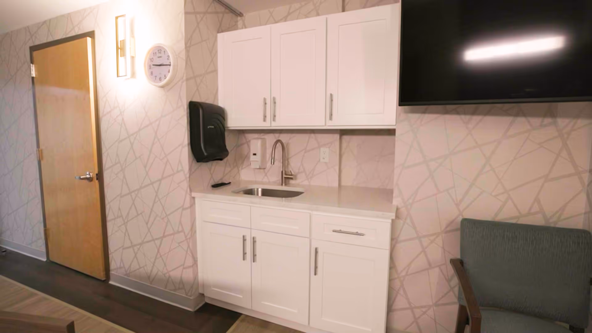 Interior view of a room with a small white kitchenette featuring a sink, white cabinets, a wall-mounted paper towel dispenser, and a soap dispenser. There is a wooden door on the left side, a wall clock above the sink, a wall-mounted light fixture, a flat-screen TV on the right, and a green upholstered chair partially visible.