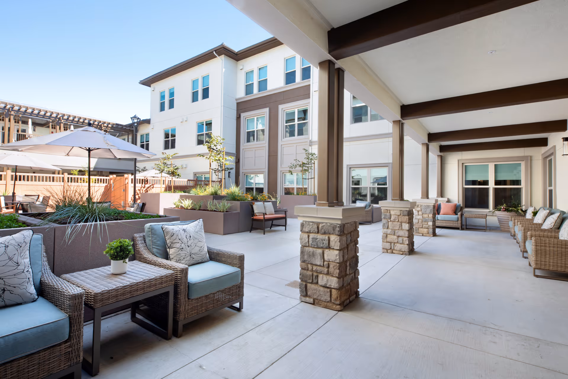 Outdoor patio area at a senior living facility with wicker chairs and cushions, small tables, potted plants, stone pillars supporting a covered walkway, and a building with multiple windows in the background under a clear blue sky.
