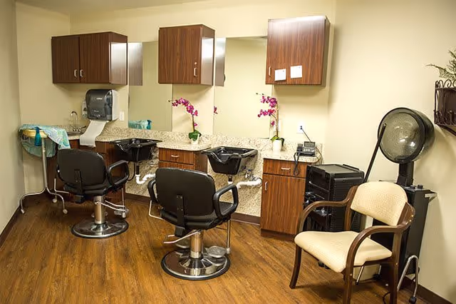 Interior view of a salon area with two black salon chairs in front of sinks and mirrors, wooden cabinets mounted on the wall, a beige cushioned chair, and a hair dryer machine on the right side. The floor is wooden and the walls are light-colored.