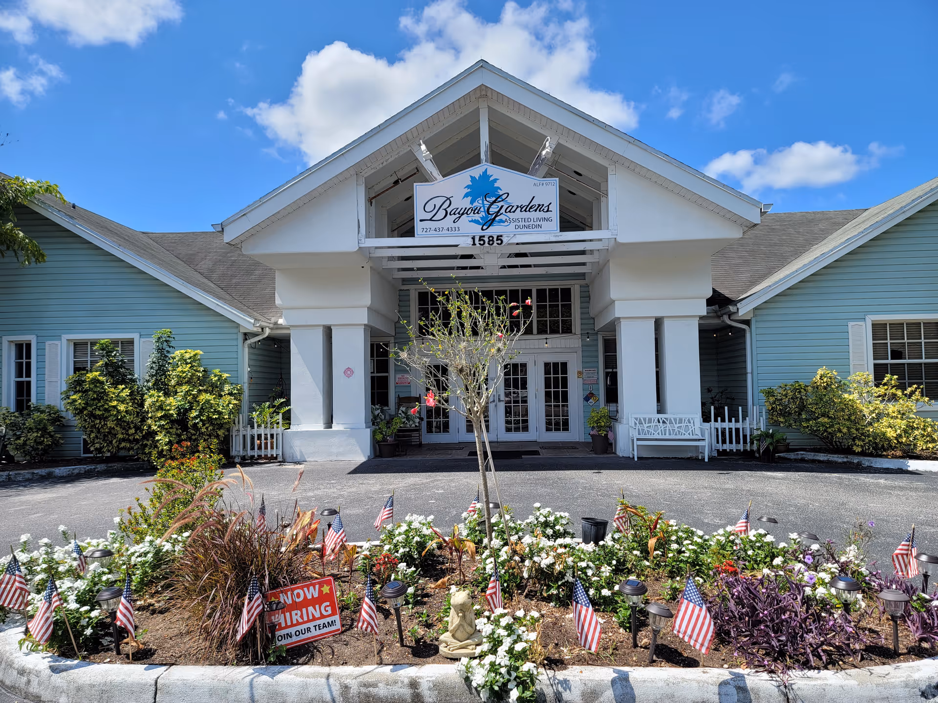 Front exterior view of Bayou Gardens Dunedin assisted living facility with a garden bed featuring flowers and small American flags in front. The building has light blue siding with white trim and a white entrance structure with a sign displaying the facility name and phone number. A 'Now Hiring Join Our Team!' sign is visible in the garden bed.